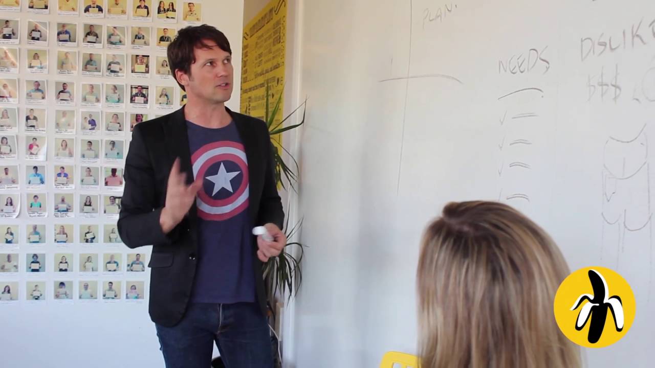 A man leading a marketing workshop, standing in front of a whiteboard talking to a woman.