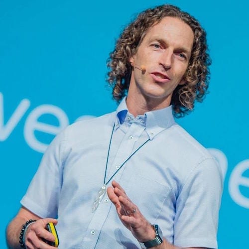 A man with curly hair standing in front of a blue background, providing mentoring on small business marketing during a marketing workshop.