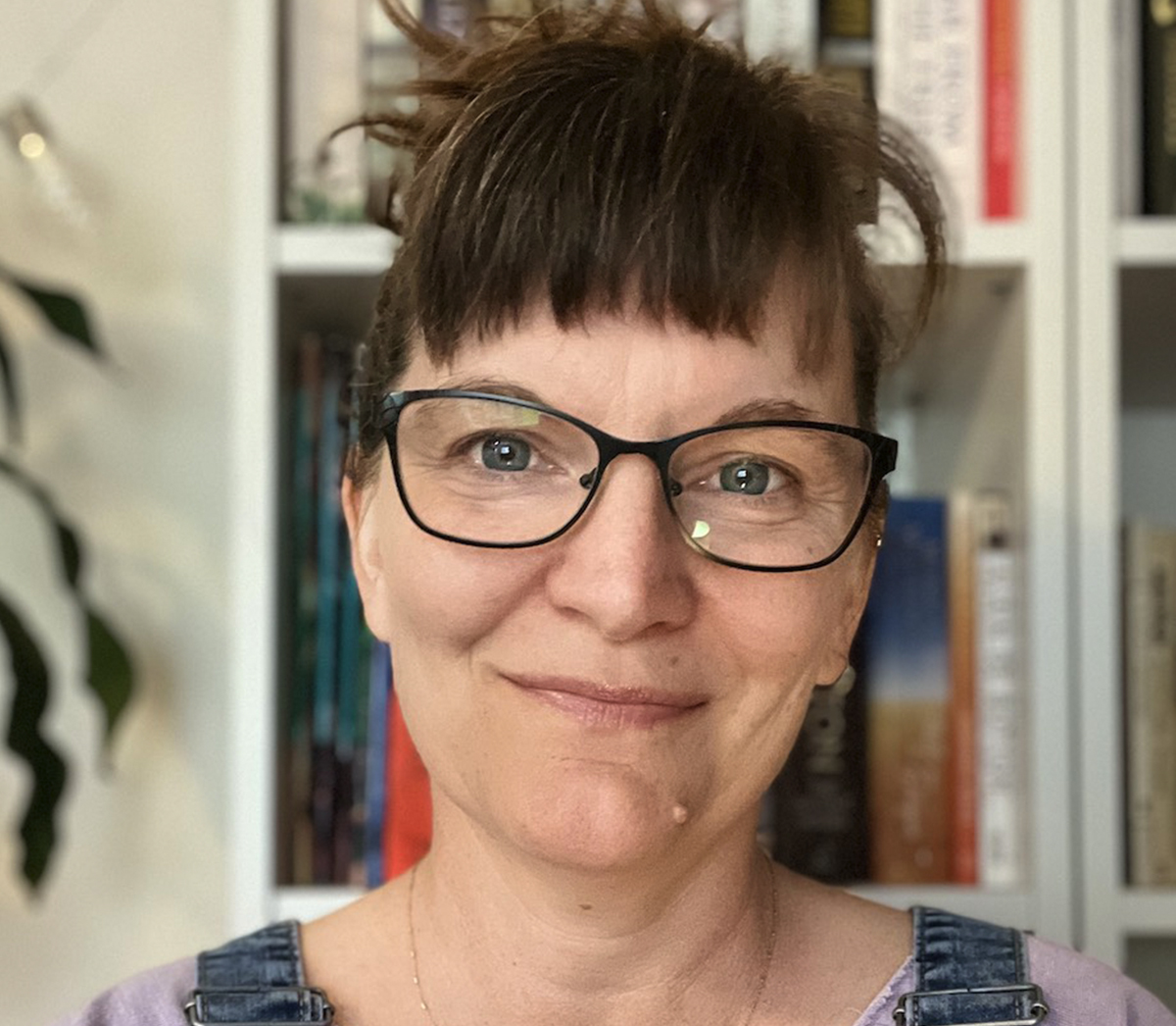 A woman in glasses is standing in front of a bookshelf.