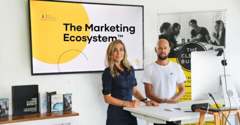 Two people stand at a desk in a modern office, presenting a slide titled "The Marketing Ecosystem™." Various books and a promotional poster are displayed in the background.