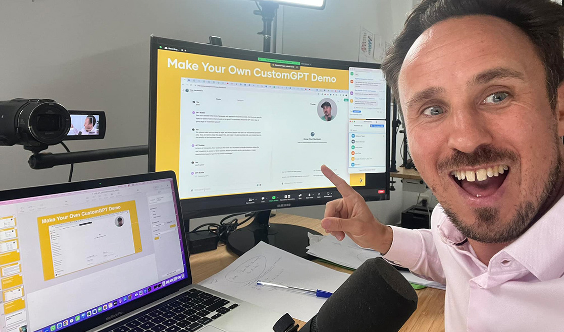 A man smiles and points at a computer monitor displaying a "Make Your Own CustomGPT Demo" presentation, with a laptop and microphone on the desk. A man smiles and points at a computer monitor displaying a "Make Your Own CustomGPT Demo" presentation, with a laptop and microphone on the desk.