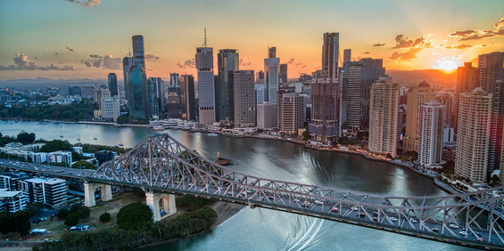 Aerial view of a city skyline with high-rise buildings, a river, and a large steel bridge at sunset.
